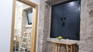 A room with a stone wall, a wooden chair, a small table with a vase, and a window with shutters.