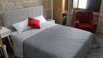 Ein modernes Hotelzimmer mit einer grau strukturierten Bettdecke, einem weißen Kissen und einem roten Akzentkissen. Es gibt einen Nachttisch aus Holz mit einer Lampe, einen roten Sessel und ein Fenster mit Blick ins Freie.