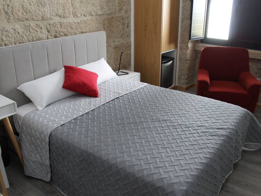 Ein modernes Hotelzimmer mit einer grau strukturierten Bettdecke, einem weißen Kissen und einem roten Akzentkissen. Es gibt einen Nachttisch aus Holz mit einer Lampe, einen roten Sessel und ein Fenster mit Blick ins Freie.