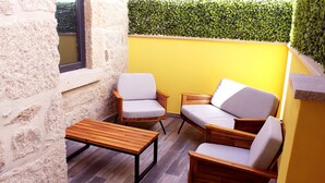 A small patio with two wooden chairs and a table, a stone wall, and a yellow wall with a green hedge.