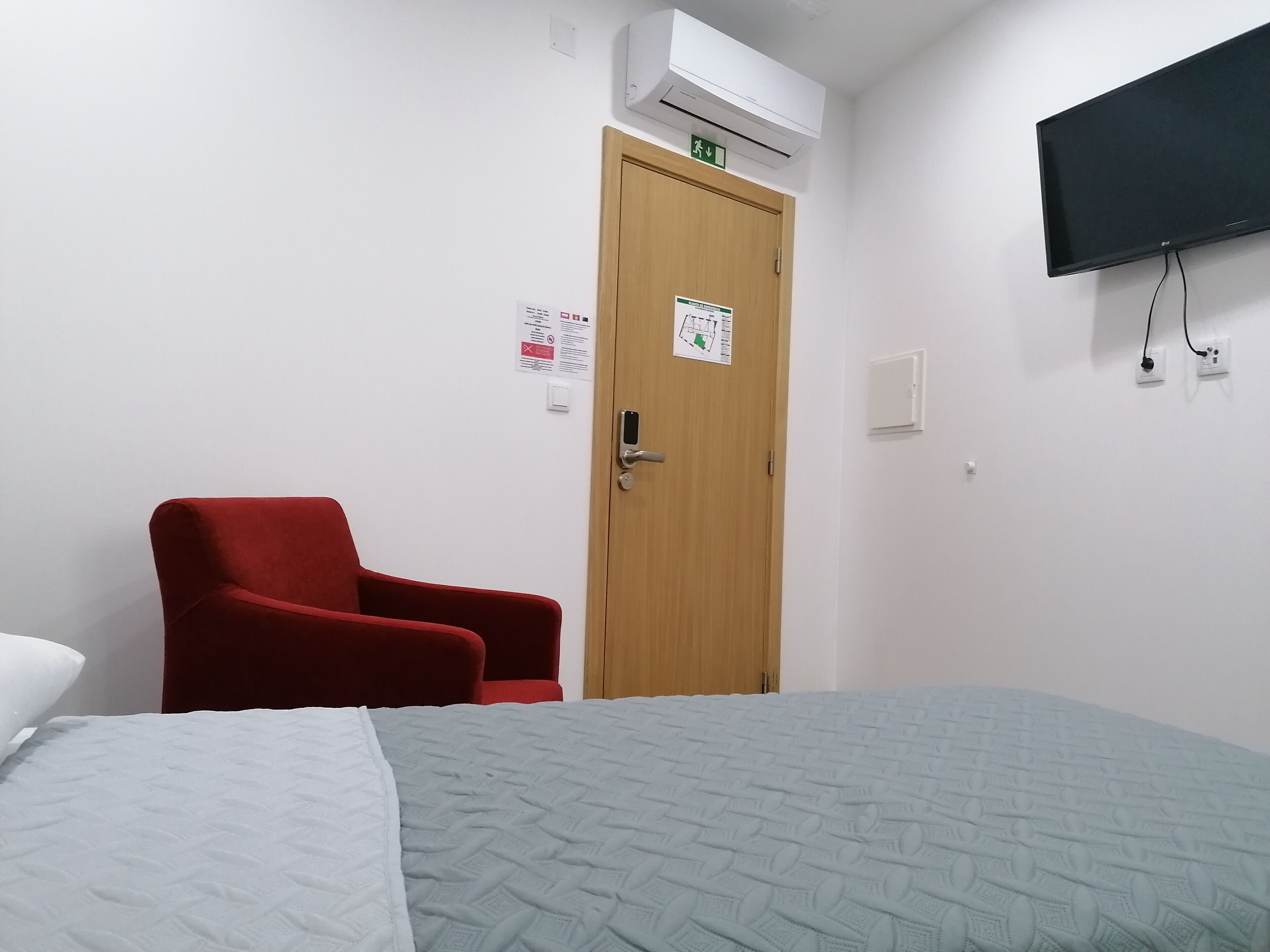Quarto de hotel com uma cama, uma poltrona vermelha, uma porta de madeira, um aparelho de ar condicionado, uma televisão e uma tomada na parede.