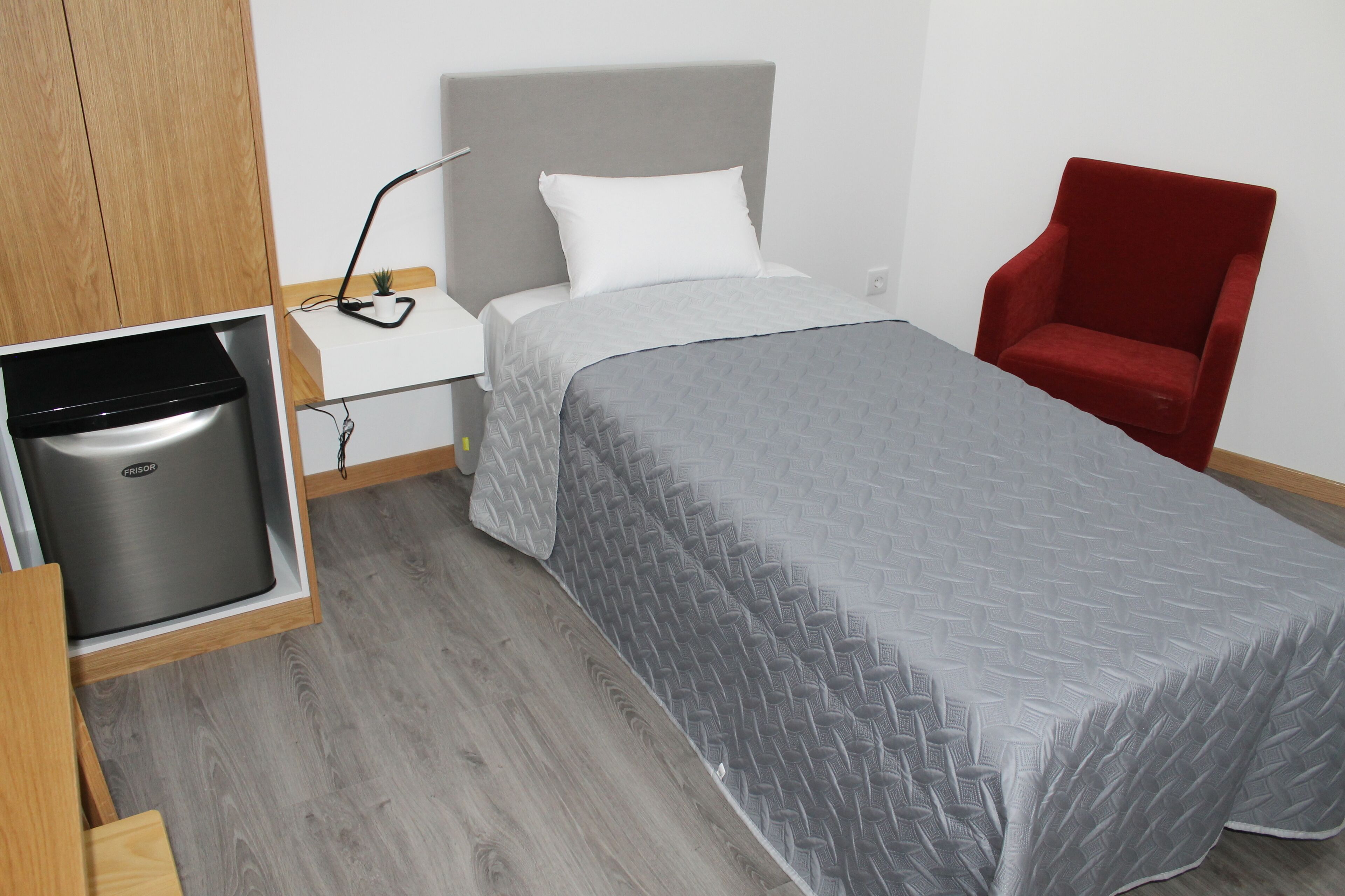 A bedroom with a bed, a red chair, a white bedside table with a lamp, and a small potted plant.