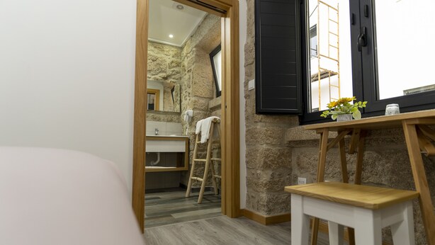 A bedroom with a bed, a wooden stool, a small table with a vase, and a view of a bathroom with a towel hanging on the wall.