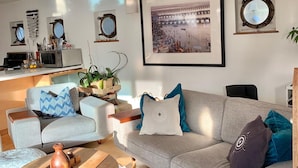 Un salon moderne avec un canapé gris, une table basse et un pouf rond. Un tableau encadré est accroché au mur, et il y a une petite fenêtre avec un miroir.