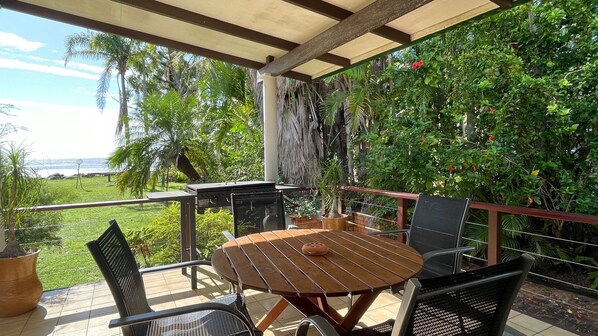 A covered patio with a wooden table and chairs, overlooking a lush garden and a view of the ocean.
