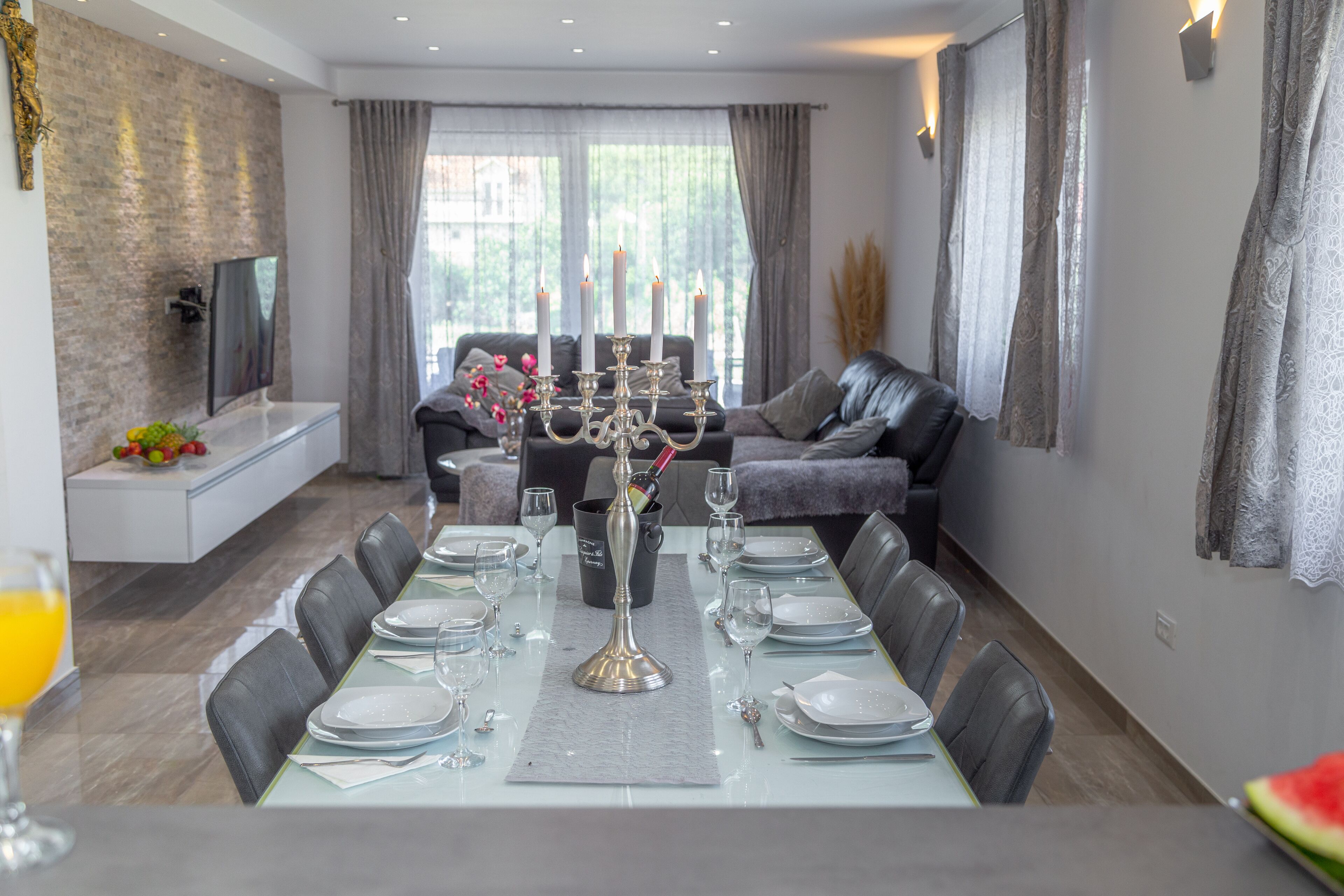 A dining area with a long table set for six, a modern kitchen in the background, and a comfortable seating area with a sofa and a TV.