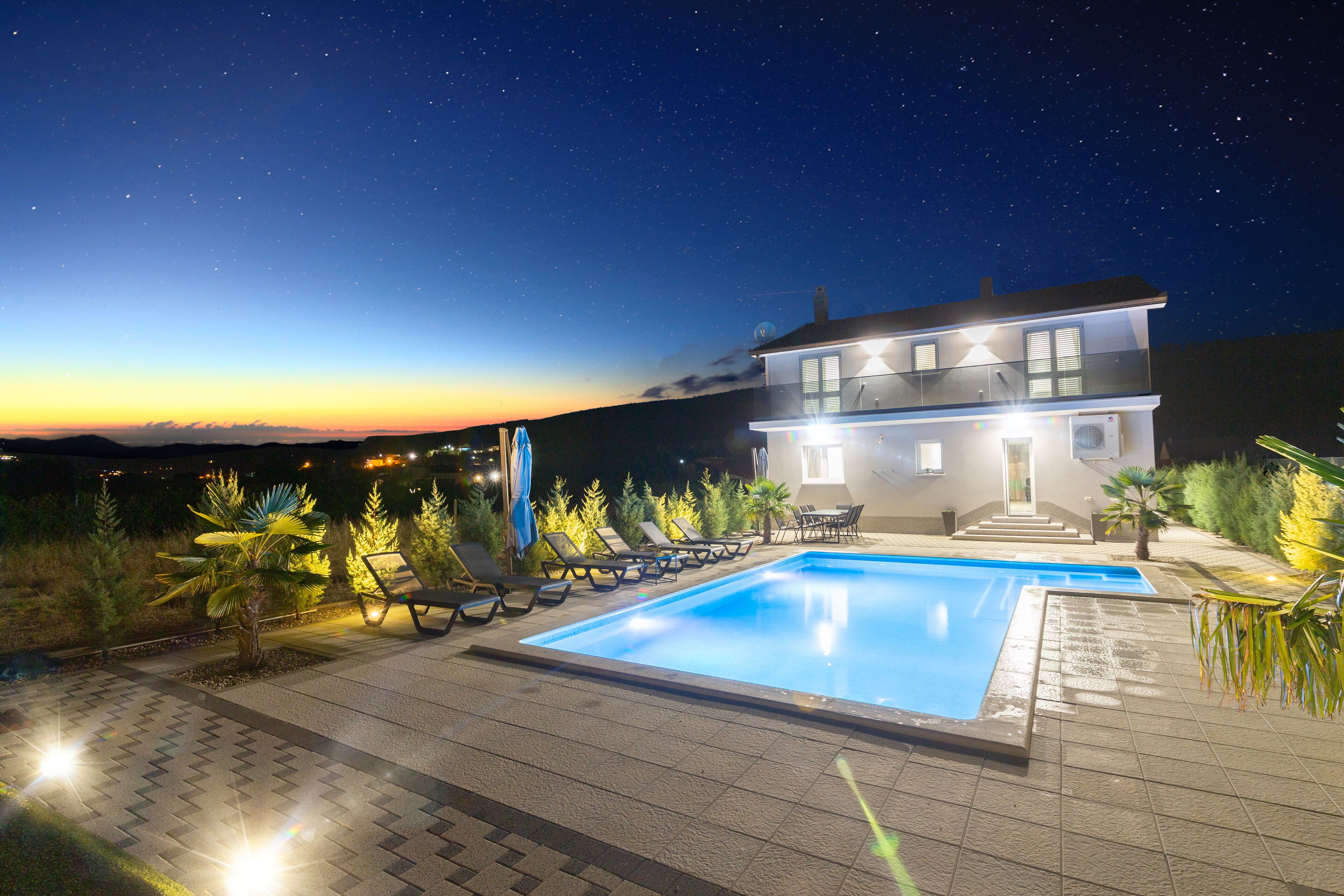 A modern two-story house with a swimming pool, surrounded by lounge chairs and palm trees.
