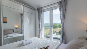Une chambre d’hôtel avec un grand lit, une table de chevet avec une bouteille et un verre, et une vue sur un balcon donnant sur un paysage.