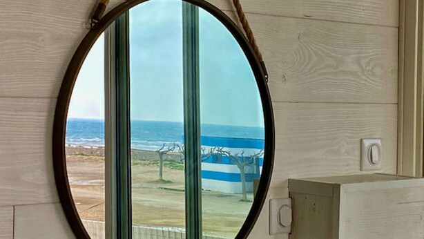 A round mirror on a wooden wall with a view of the ocean and a blue and white building.