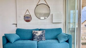 A teal sofa with a tropical patterned pillow, a round mirror, and a small framed picture on a white wall.
