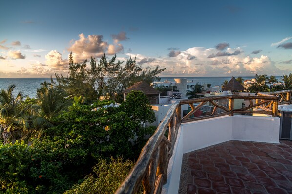 A rooftop terrace with a view of the sea, surrounded by tropical vegetation and buildings with thatched roofs. - Awesome 2 BDR apartment, safe, clean, WiFi, and only steps from the beach. (Puerto Morelos)