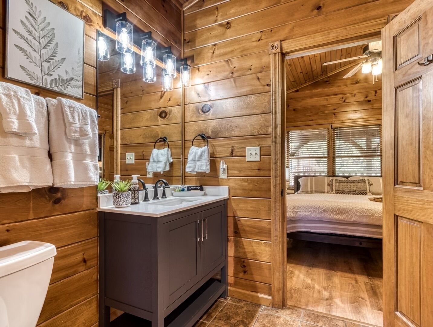 A wooden cabin bathroom with a vanity, towels, and a bed visible through an open door.