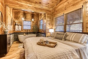 A log cabin-style bedroom with a large bed, a wooden dresser, a vanity, and a bathtub. - Luxurious cabin with an amazing wooded view (Gatlinburg)