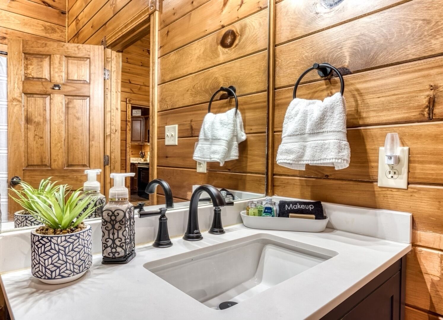Un baño con paredes de madera, un lavamanos blanco, grifos negros, un espejo y toallas colgadas en ganchos.