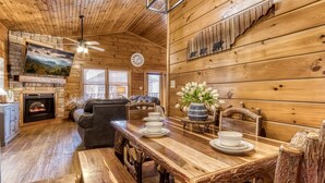 Un interior acogedor de cabaña con una mesa de comedor de madera para cuatro, una chimenea, un sofá y un televisor montado en la pared.