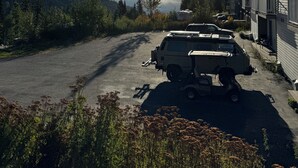 Un'area di parcheggio con un veicolo, un edificio con balconi e un paesaggio montano sullo sfondo.