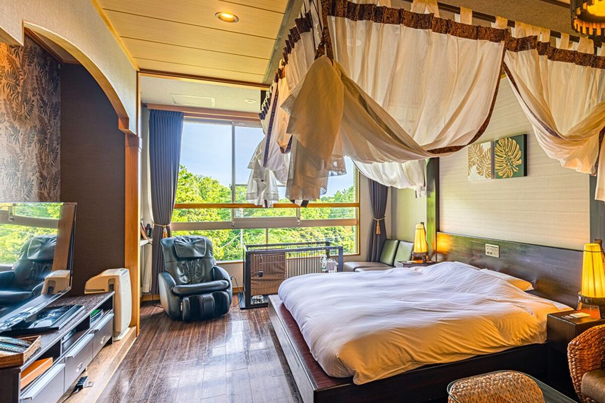 A spacious bedroom with a large bed, a massage chair, a TV, and a balcony with a view of greenery.