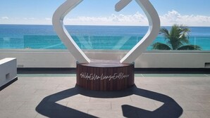 A modern, minimalist sculpture with a wooden base and a white, geometric design, set against a backdrop of a clear blue sky and ocean. - Direct Beachfront Paradise: Studio w/ Balcony + Resort Amenities (Cancún)
