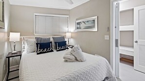 Une chambre bien rangée, avec un lit, des oreillers, une table de chevet, un ventilateur de plafond, une fenêtre avec des stores, un tableau encadré et une porte donnant sur un placard.