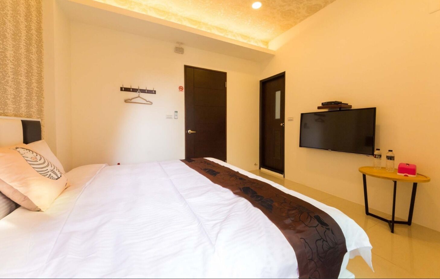 A hotel room with a bed, a television, a small table with a water bottle, and a coat rack.