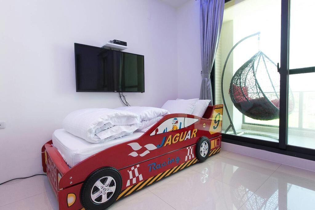 Una cama con diseño de coche de carreras, un televisor de pantalla plana montado en la pared y un sillón colgante junto a la ventana.