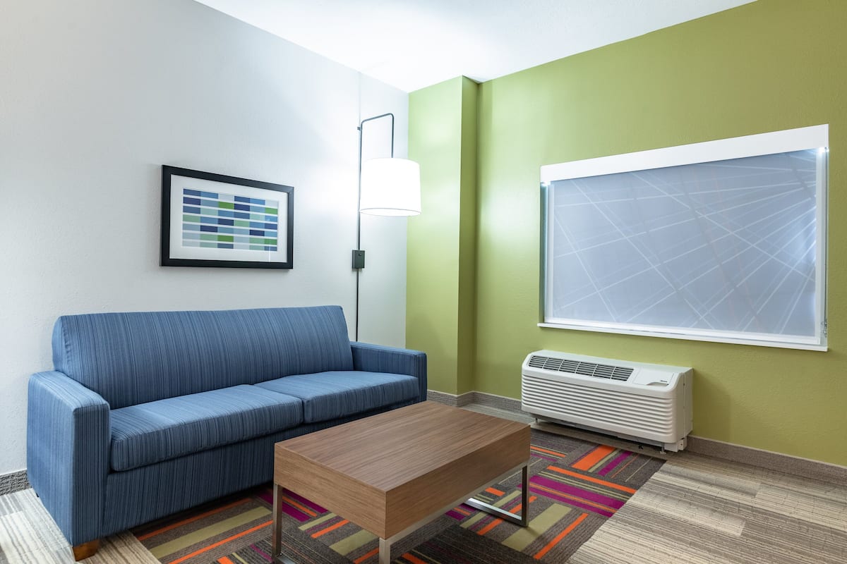 A room with a blue sofa, a wooden coffee table, a colorful rug, a framed picture on the wall, a floor lamp, and a large window with blinds.