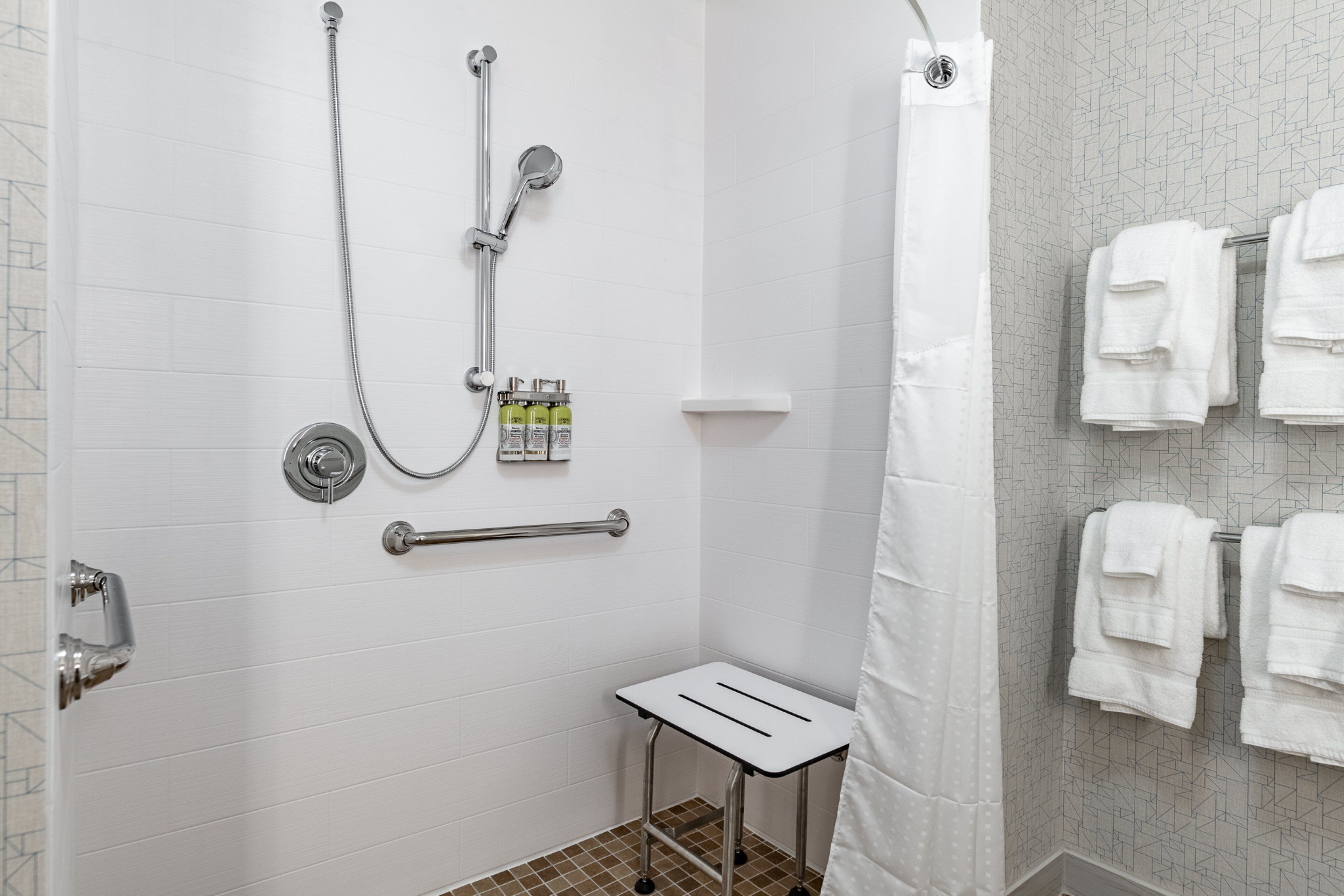 Douchegedeelte met een handdouche, een wandmengkraan en een klein plankje met toiletartikelen. Er is een handgreep aan de muur en een witte handdoekhouder met opgevouwen handdoeken.