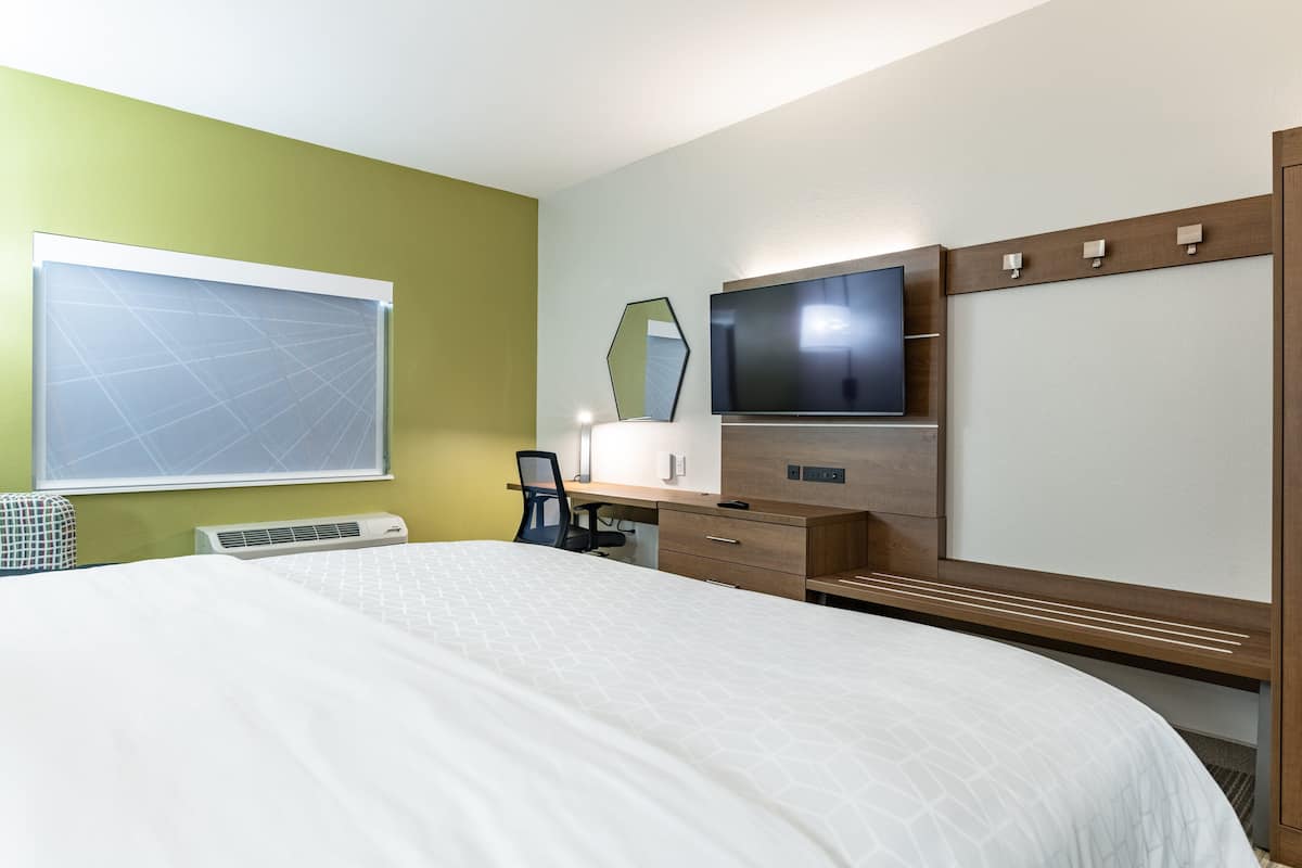 A hotel room with a large bed, a desk with a chair, a flat-screen TV, a mirror, and a window with blinds.