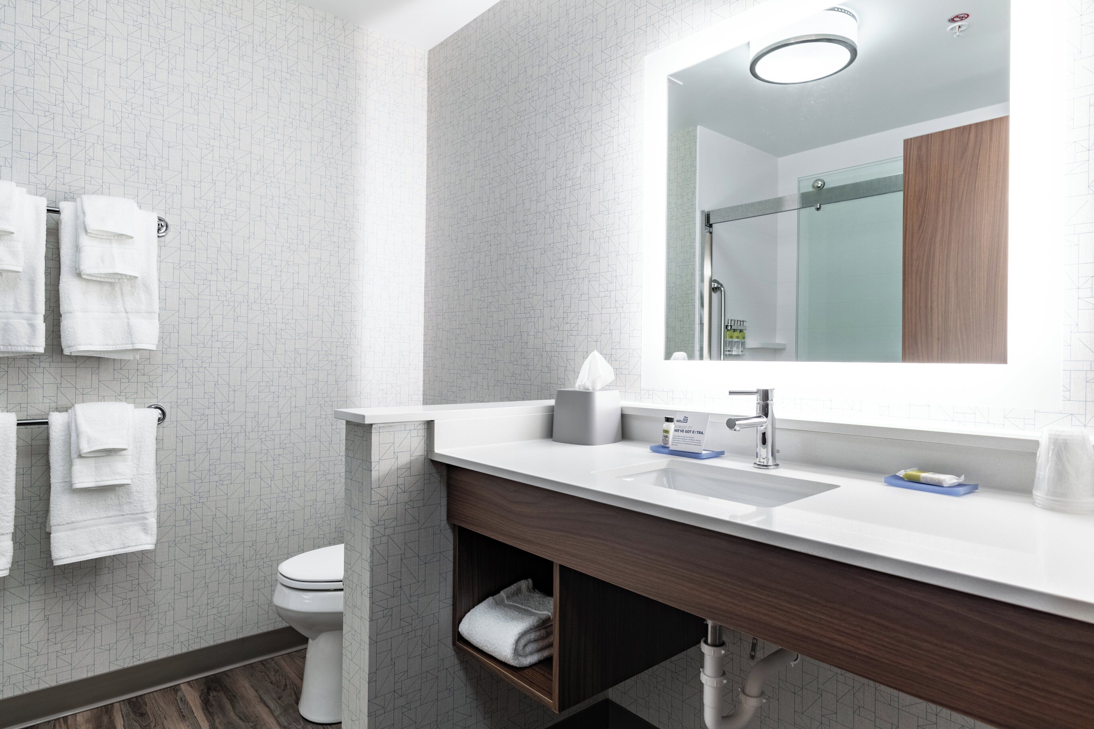 A modern bathroom with a white sink, a mirror, and a toilet. There are towels hanging on a rack and a shelf under the sink.