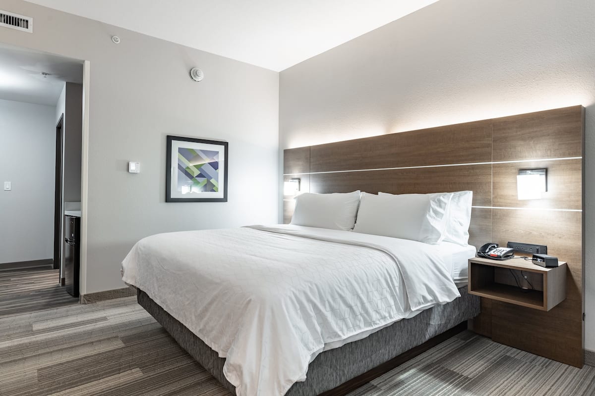 A modern hotel room with a large bed, a bedside table with a phone and charger, a framed picture on the wall, and a built-in shelf.