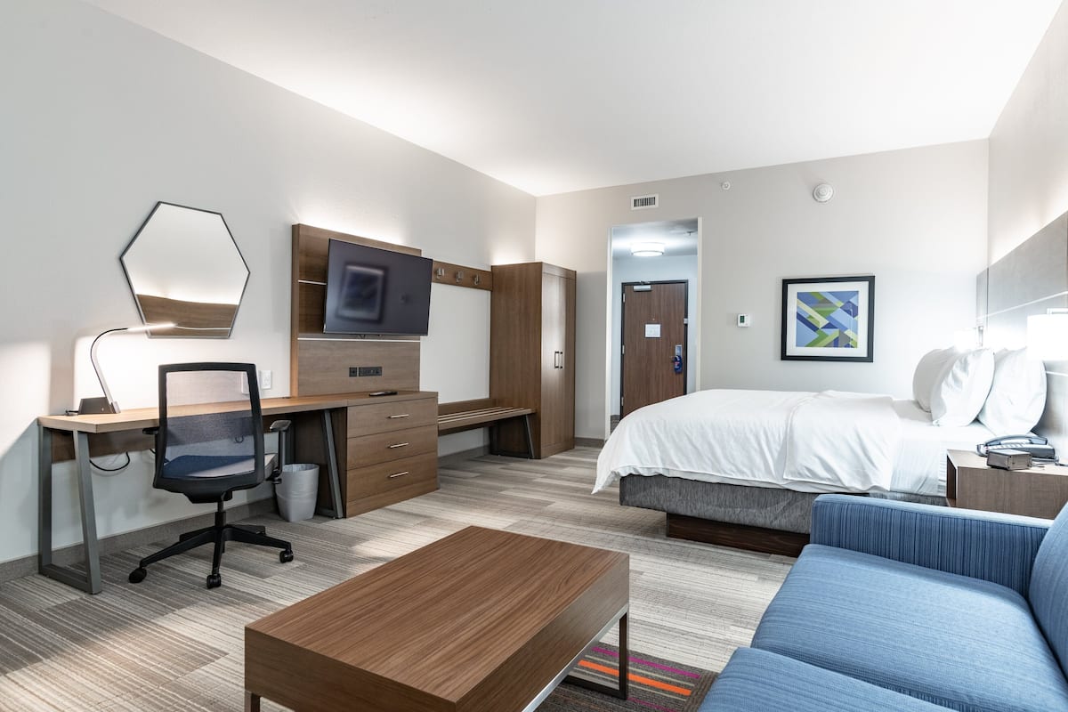 A hotel room with a bed, a desk with a chair, a TV, a sofa, and a small table.