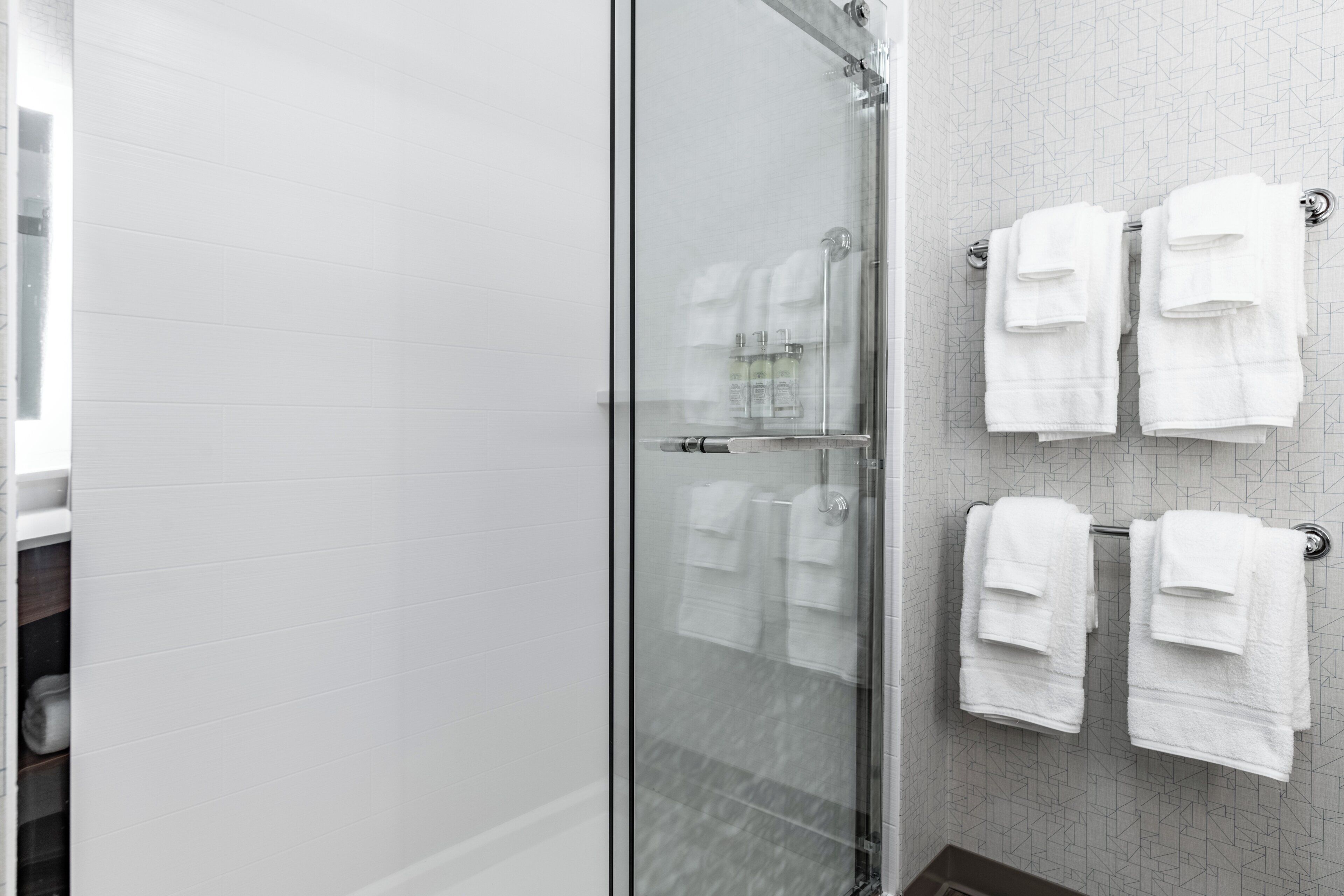 Un baño moderno con cabina de ducha de vidrio, paredes de azulejos blancos y toallas blancas ordenadas en ganchos.