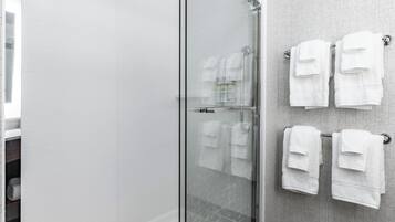 Un baño moderno con cabina de ducha de vidrio, paredes de azulejos blancos y toallas blancas ordenadas en ganchos.