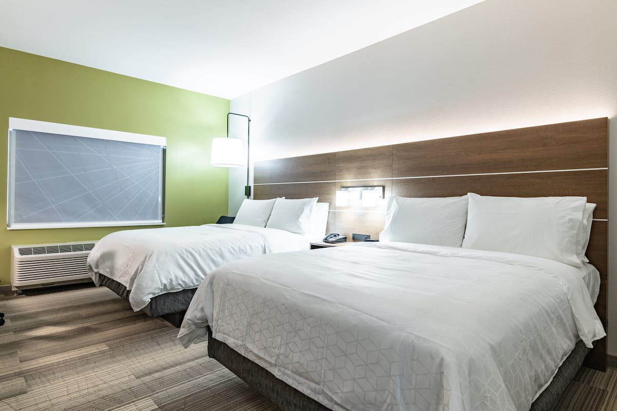 A hotel room with two beds, a large window, a wall-mounted air conditioner, and a bedside table with a lamp.