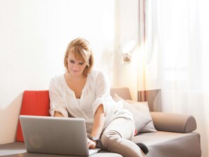 A person sitting on a sofa with a laptop, a red pillow, and a grey cushion. - Novotel Duesseldorf Airport (Düsseldorf)