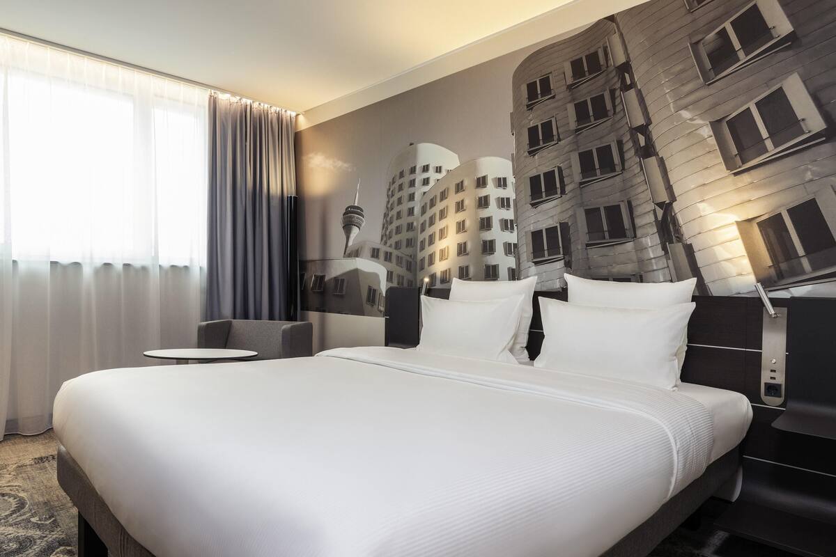 Ein Hotelzimmer mit einem großen Bett, einem kleinen runden Tisch und einer Stadtlandschaftsmalerei an der Wand.