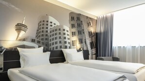 Une chambre d’hôtel avec deux lits, une fresque représentant un paysage urbain sur le mur, une lampe et une fenêtre avec des rideaux.