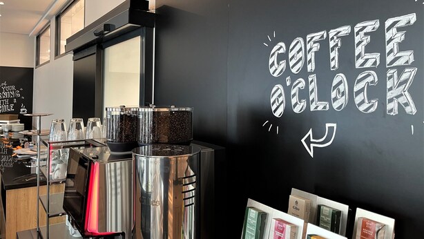L’intérieur d’un café, avec un tableau noir sur lequel on peut lire « COFFEE O’CLOCK » et une machine à café.