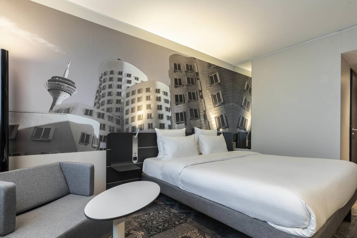 Ein Hotelzimmer mit einem großen Bett, einem kleinen runden Tisch, einer Couch und einem Wandbild von Gebäuden.