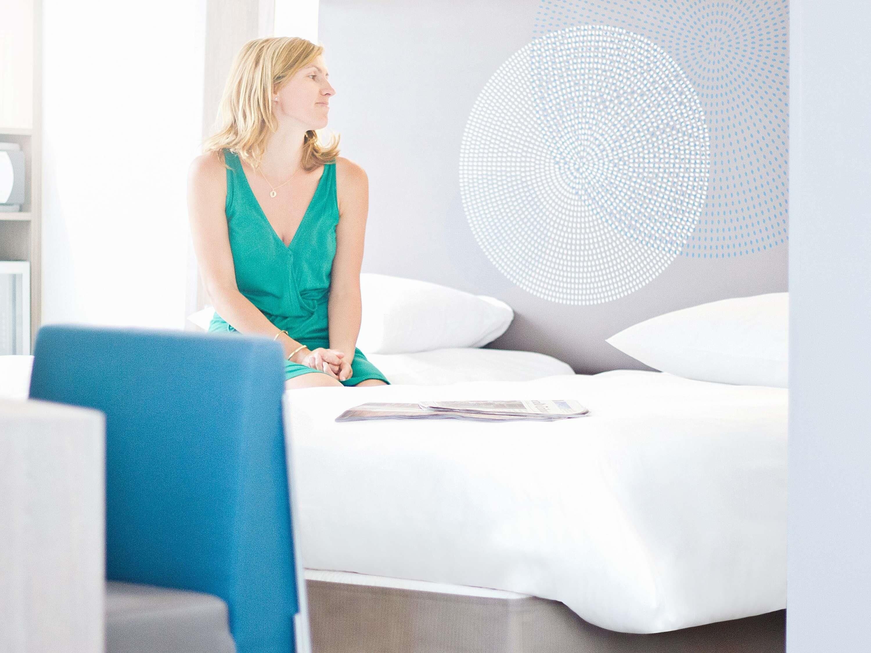 A woman sitting on a bed in a room with a modern design, including a circular light fixture on the wall.