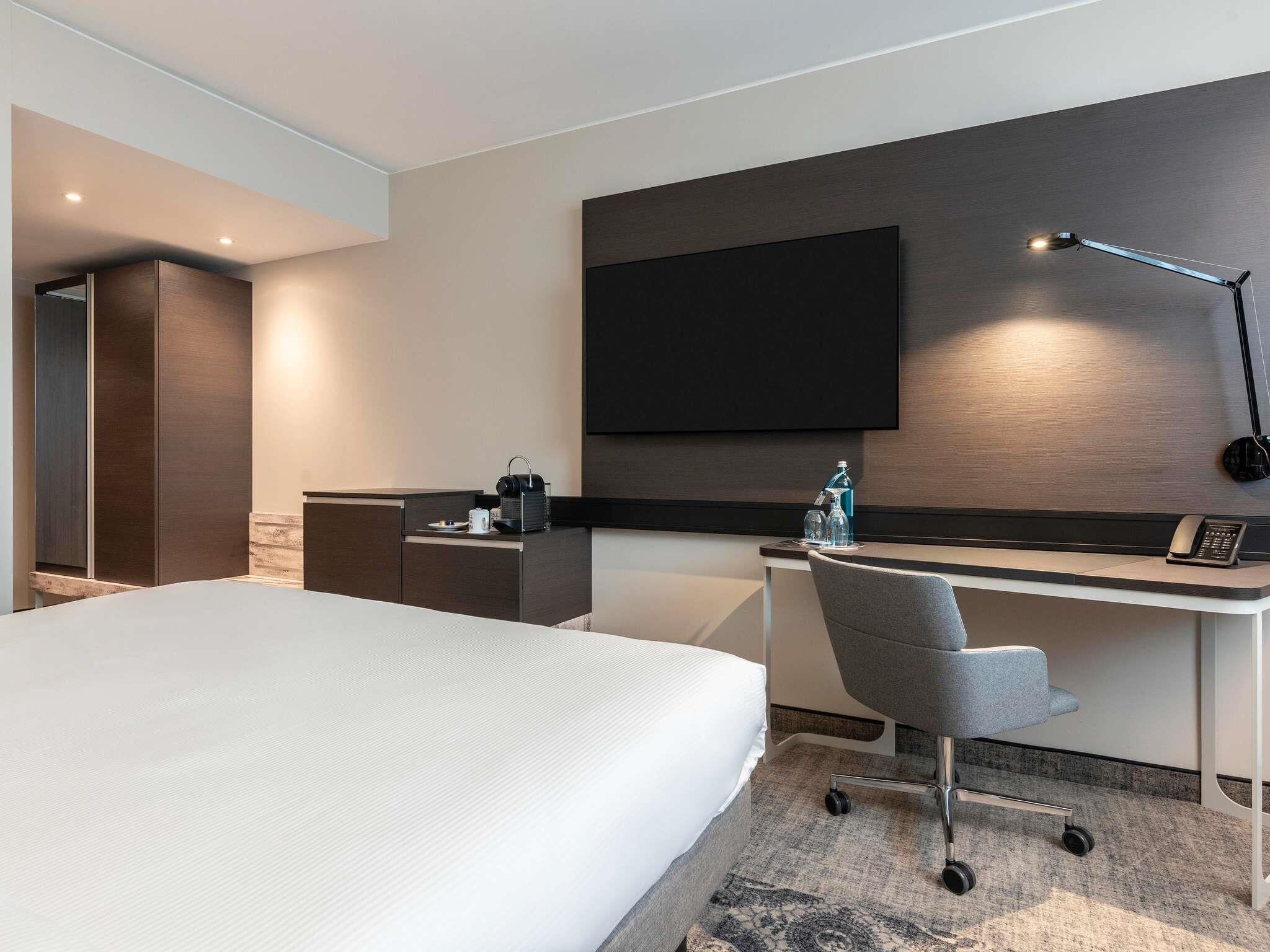 A modern hotel room with a large bed, a desk with a chair, a flat-screen TV mounted on the wall, and a desk lamp.