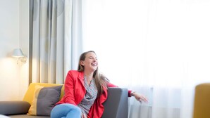 Eine Person sitzt auf einem Sofa mit einer Zeitschrift, einer Lampe und einem Fenster mit Vorhängen.