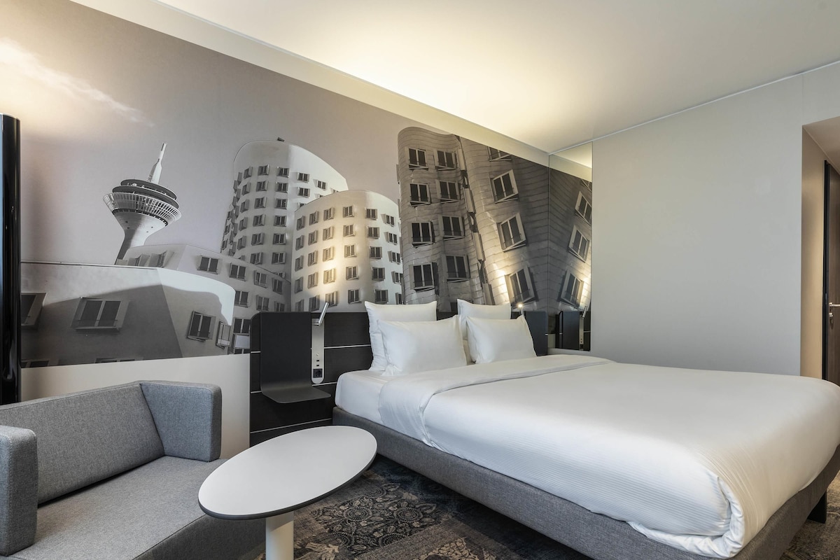 Een hotelkamer met een groot bed, een klein rond tafeltje, een bank en een muurschildering van gebouwen.