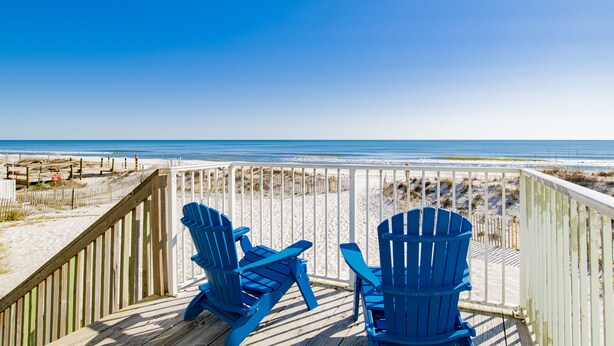 Terraza de madera con dos sillas Adirondack azules con vista a una playa.