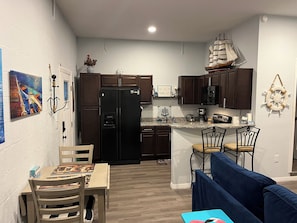 Eine kompakte Küche mit dunklen Holzschränken, einem schwarzen Kühlschrank und einer kleinen Kochinsel mit Spüle und Mikrowelle. Im Wohnbereich steht ein Esstisch mit Stühlen sowie eine blaue Couch.