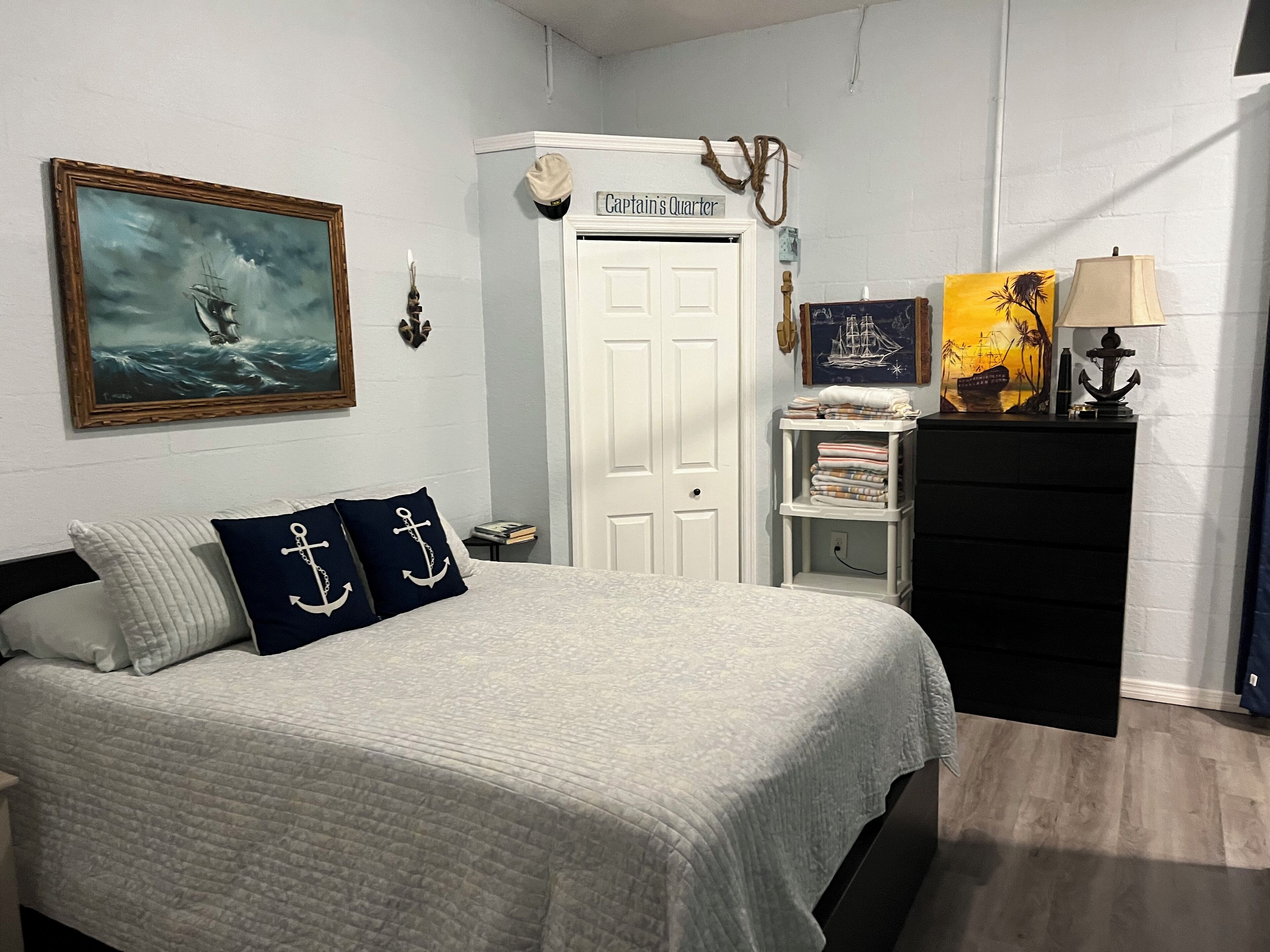 A bedroom with a bed, a painting of a ship, a door labeled "Captain's Quarters", and a small shelf with books and a lamp.