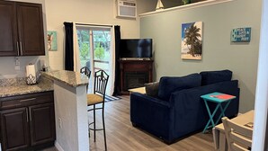 A living room with a dark blue sofa, a small table, and a TV. There is a kitchen area with dark cabinets and a granite countertop.