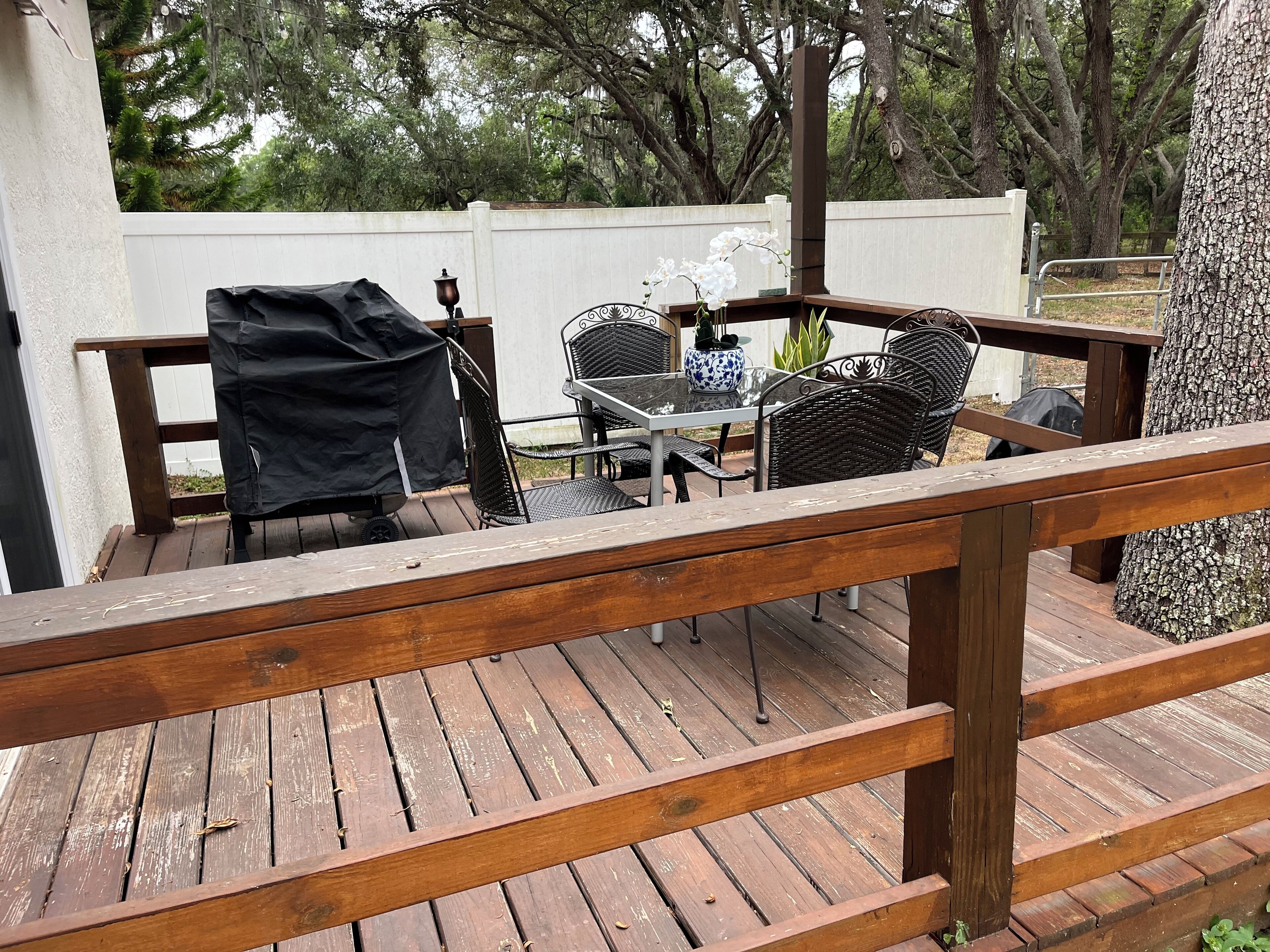 A wooden deck with a table and chairs, surrounded by trees and a white fence.