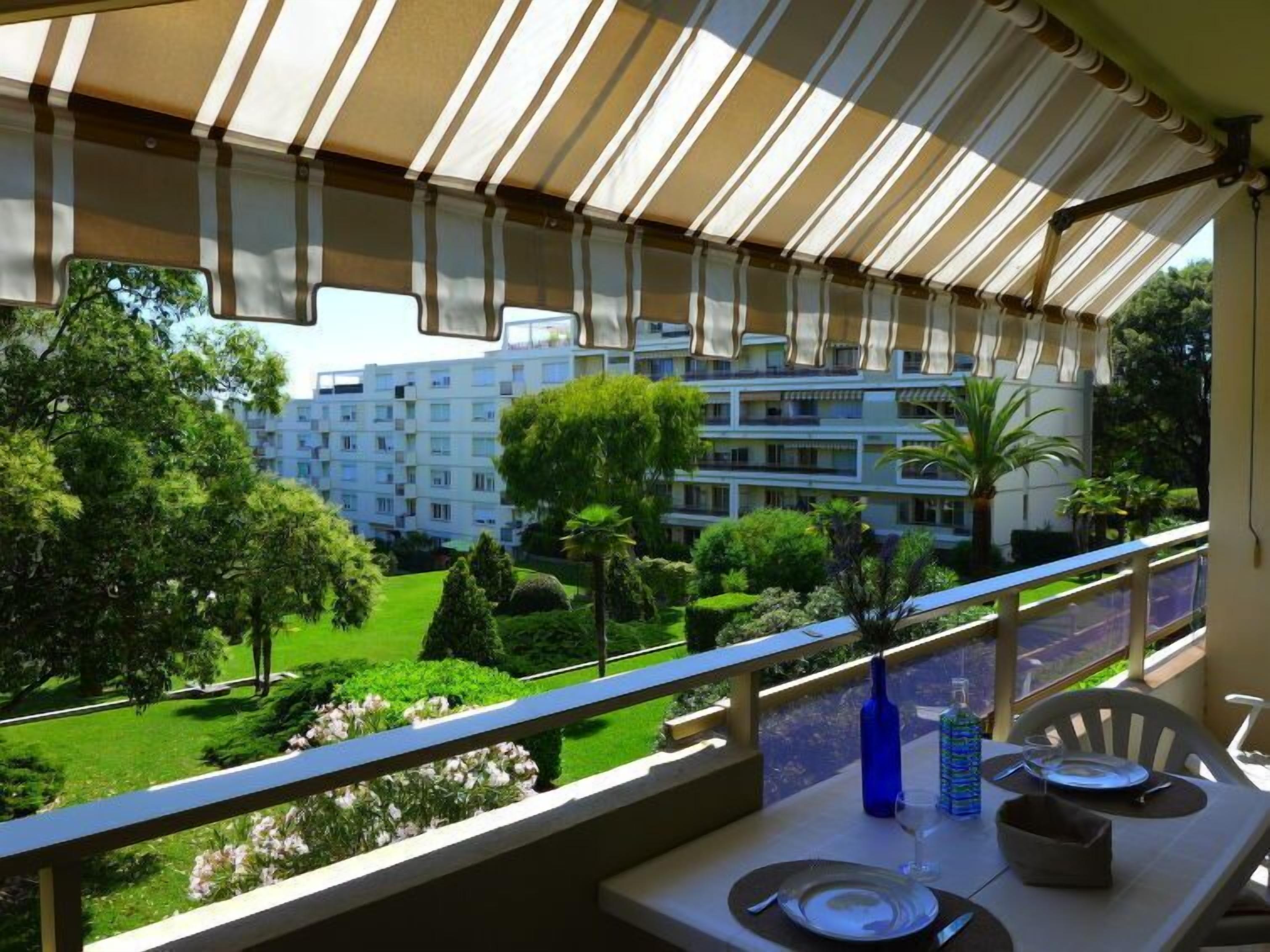 Un balcón con una mesa lista para comer, con vistas a un jardín cuidado y un edificio de varios pisos.