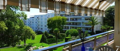 A balcony with a table set for dining, overlooking a well-maintained garden and a multi-story building.